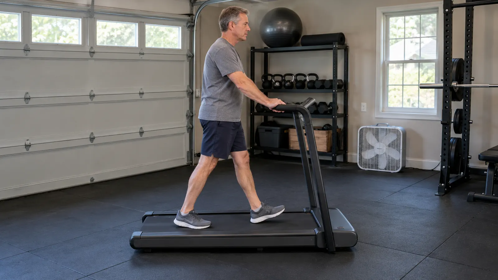 A man in his fifties walking on a walking pad treadmill, hands resting on the handle bar, garage setting with rubber stall mats