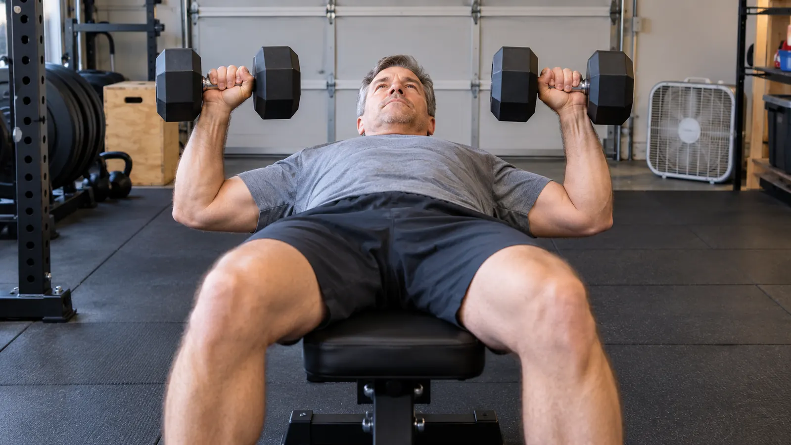 A man in his fifties doing dumbbell presses on an adjustable weight bench in a home garage