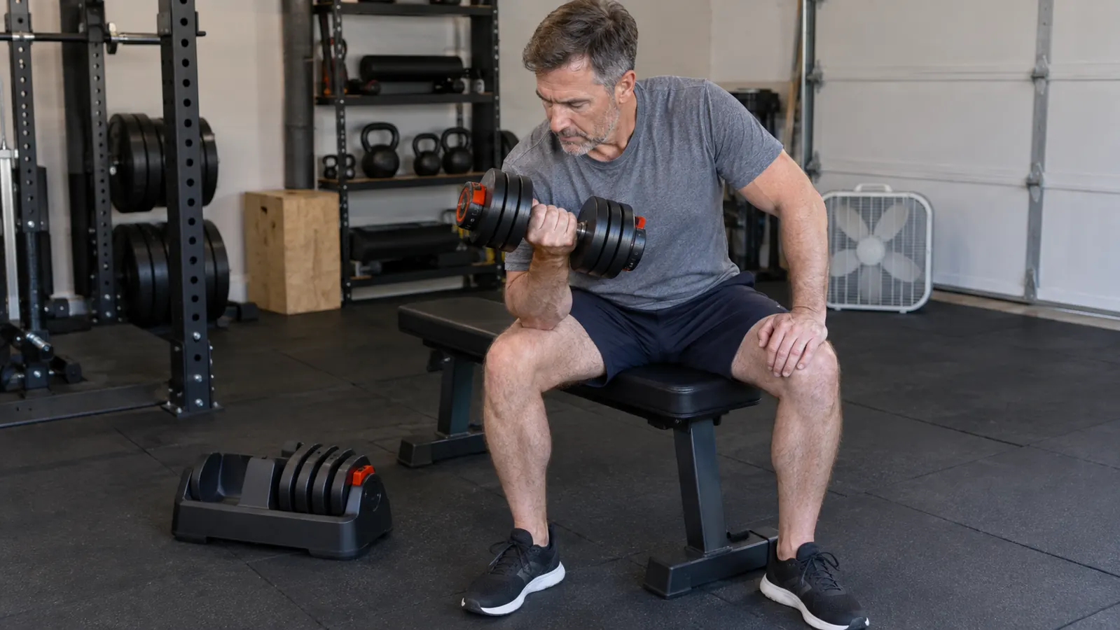 A man in his fifties seated on a workout bench, holding an adjustable dumbbell at the top of a curl while the unused plates stay in the base beside him