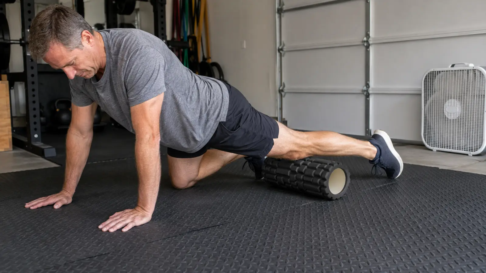 A person kneeling on a mat, rolling a calf over a foam roller on a rubber garage floor