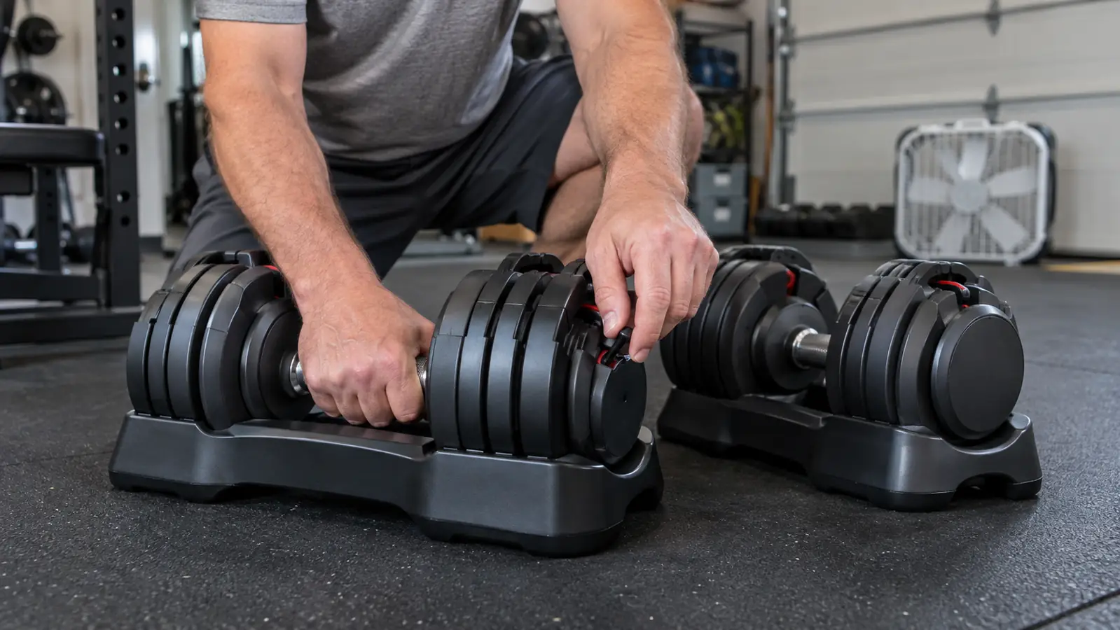 A hand sliding the selector pin on the side of an adjustable dumbbell resting on a rubber garage mat