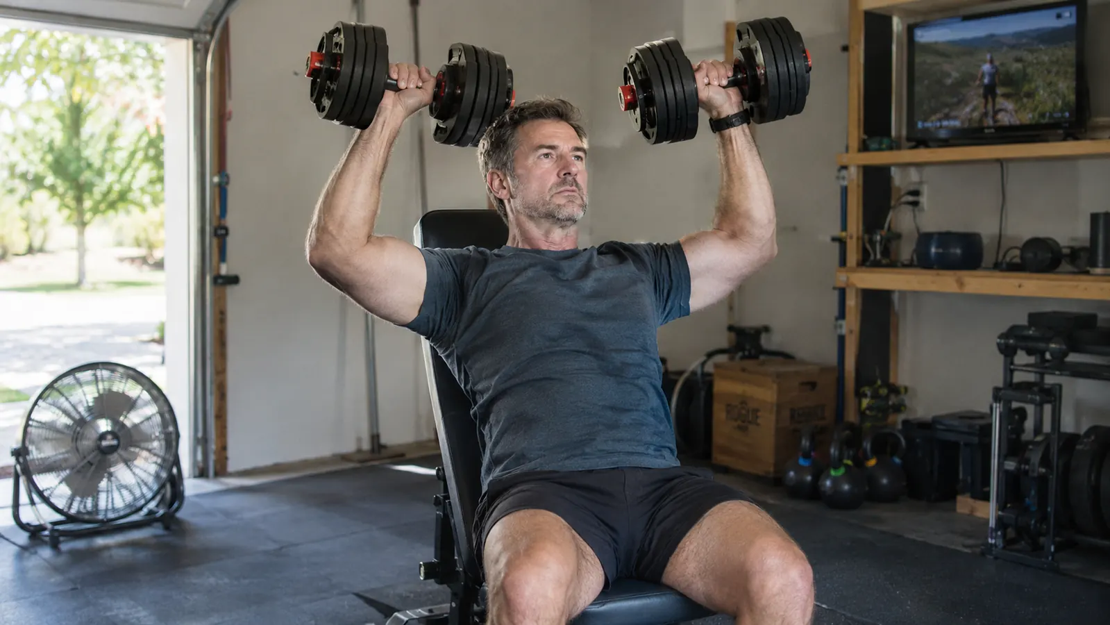 A man in his fifties doing a seated shoulder press with a pair of adjustable dumbbells on a weight bench in a garage gym