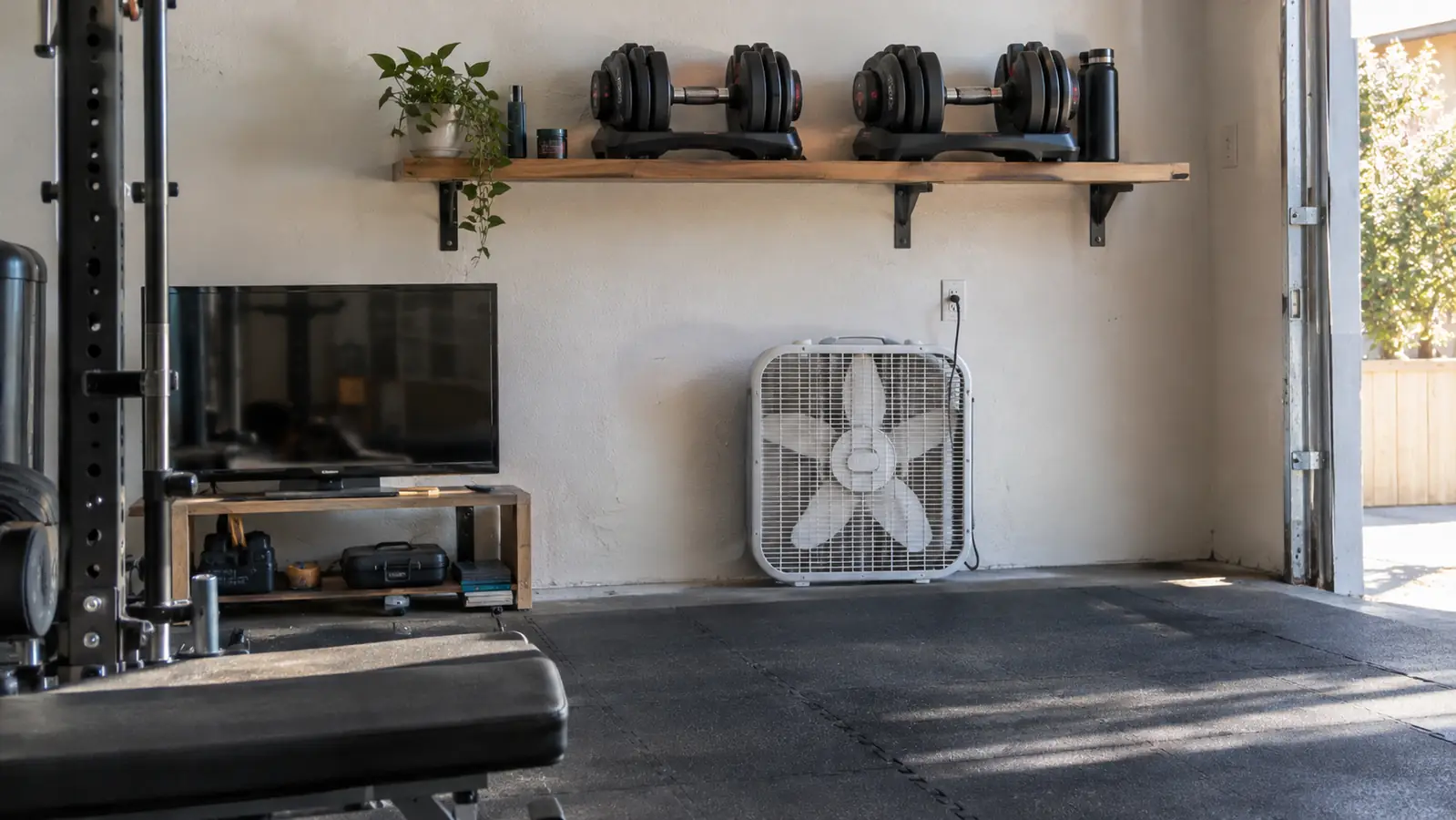 A pair of adjustable dumbbells stored on a wooden shelf above a box fan in a home garage