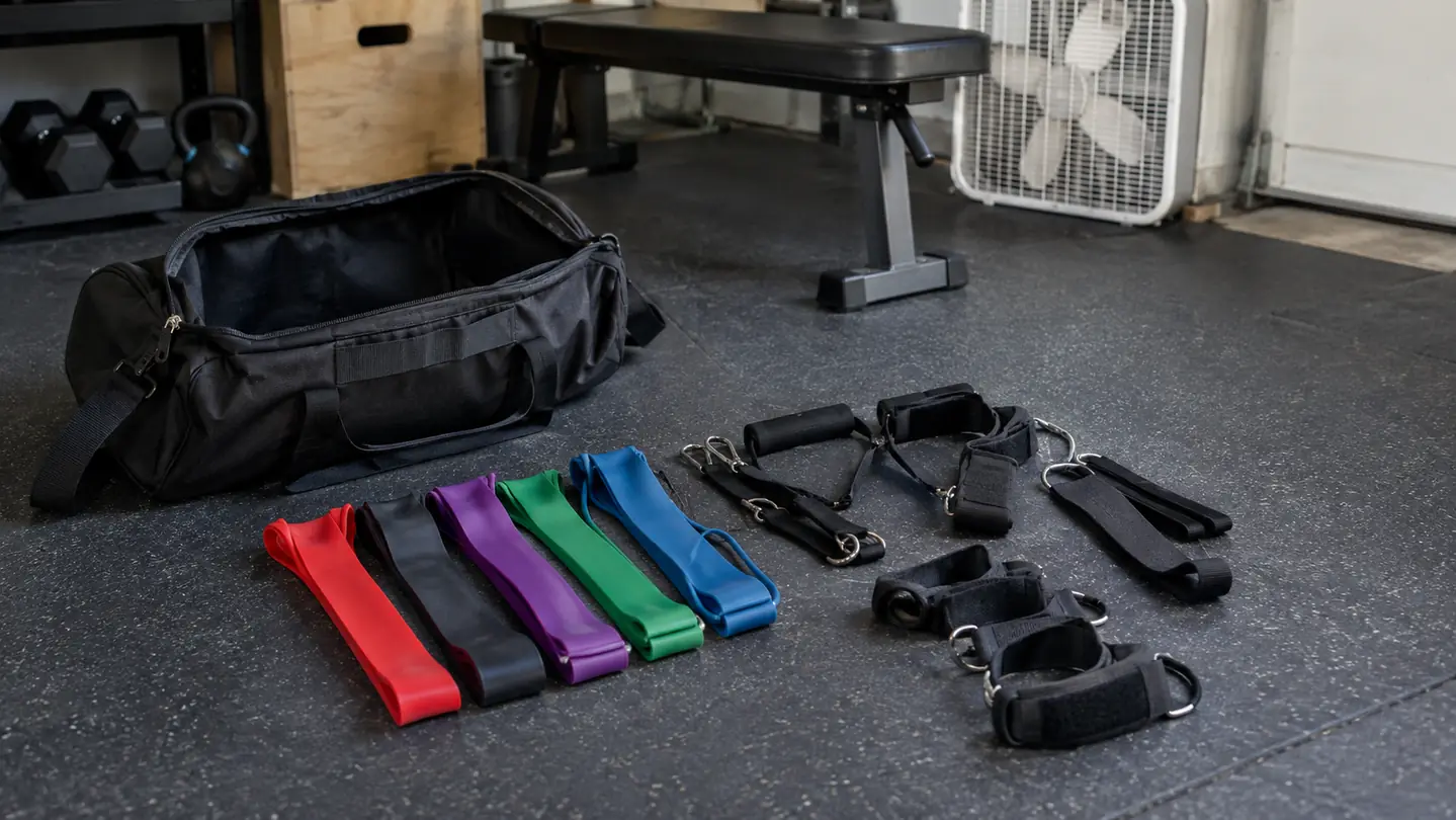 A zippered carry bag open on a rubber stall mat with resistance bands, handles, and ankle straps laid out neatly beside it