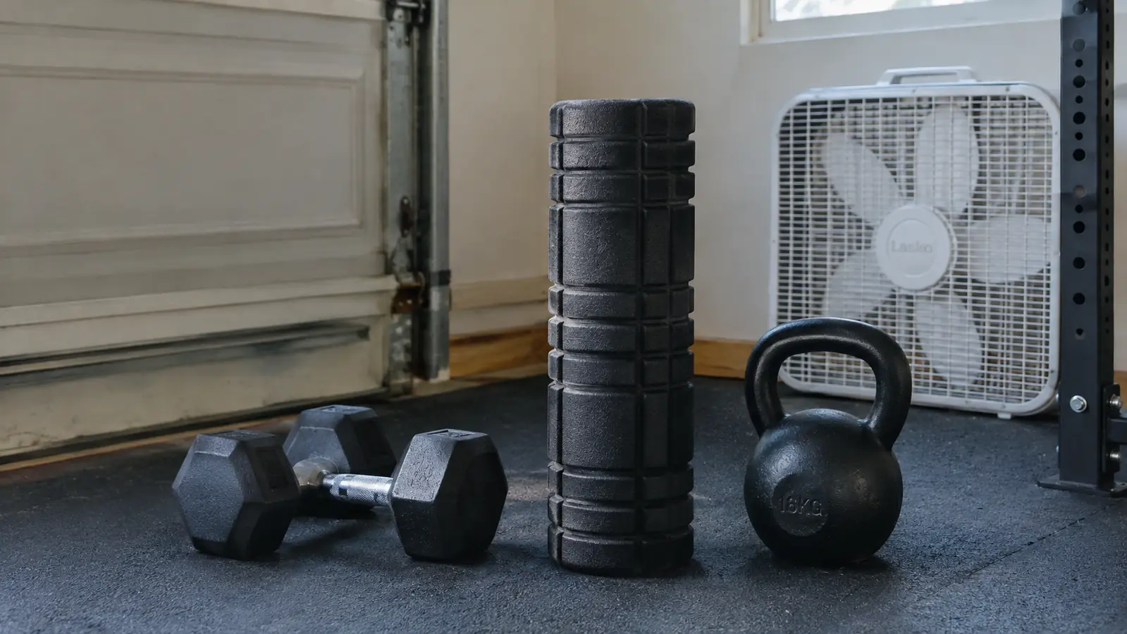 A foam roller standing upright on a garage floor next to a pair of dumbbells and a kettlebell